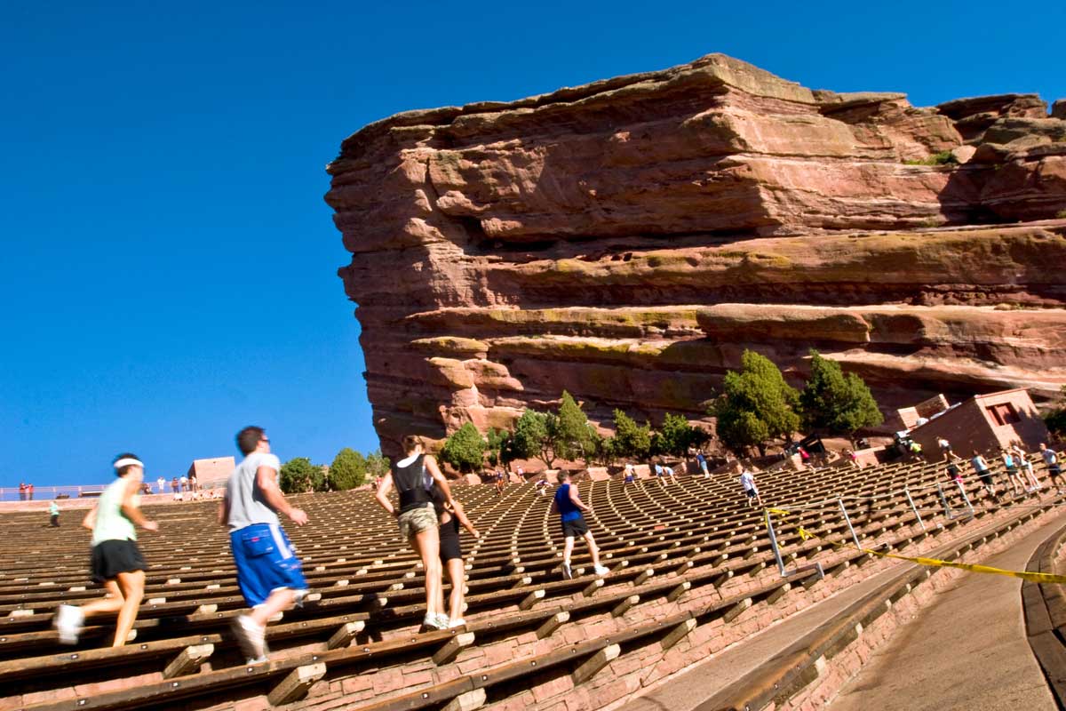 People run the steps at Red Rocks. The giant geologic red-rock formation sits against a bright-blue sky in the distance.