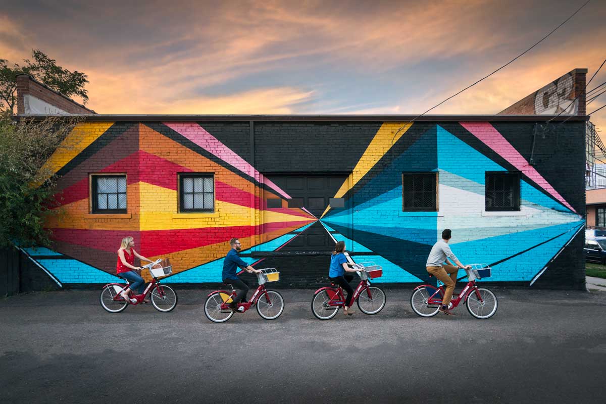 Four people on bikes ride past geometric wall art on a mural tour in Denver's RiNo neighborhood