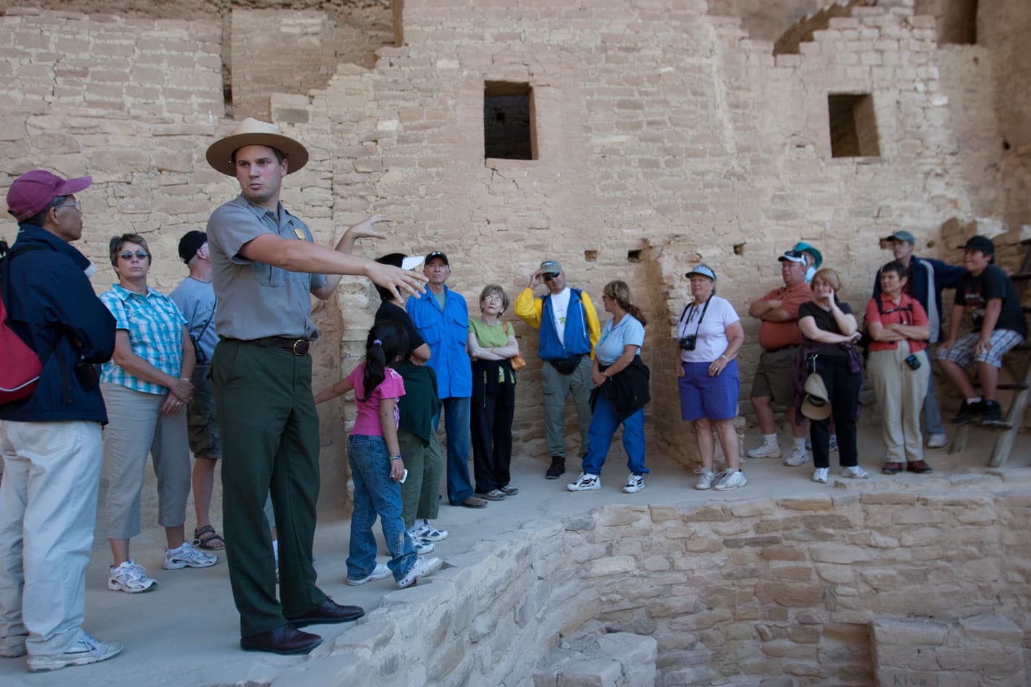 A national park ranger leads a tour of many people standing above a stone cliff dwelling basement with cliff dwelling walls with windows behind the group.