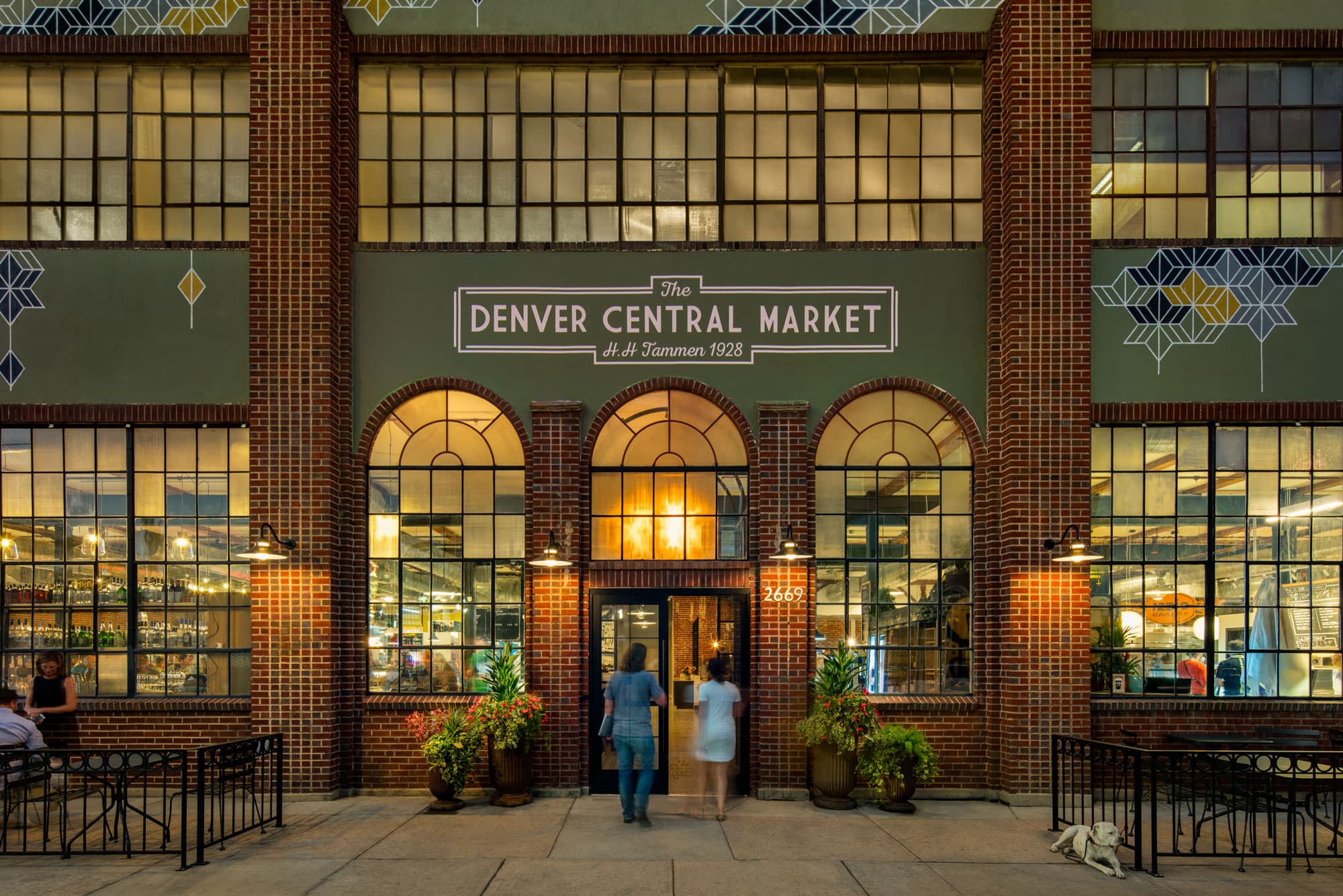 Two people walk towards the glass doors of a red-brick building with the number 2669 lit up beneath a lamp and a sign that says, "The Denver Central Market."