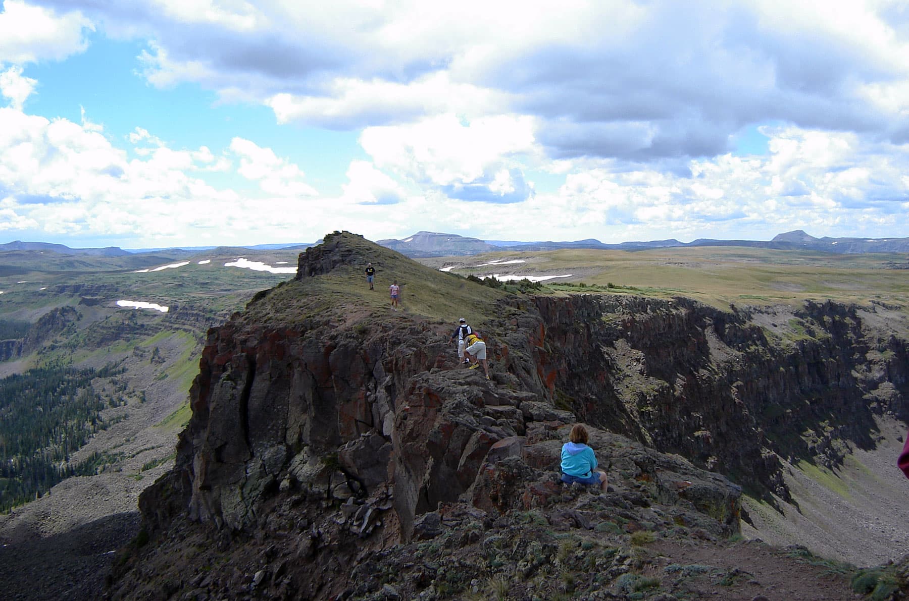 Five people hike Devil's Causeway's narrow trail, rocky gray trail on a cloud-filled summer's day. In the background white snow can be seen in spots.