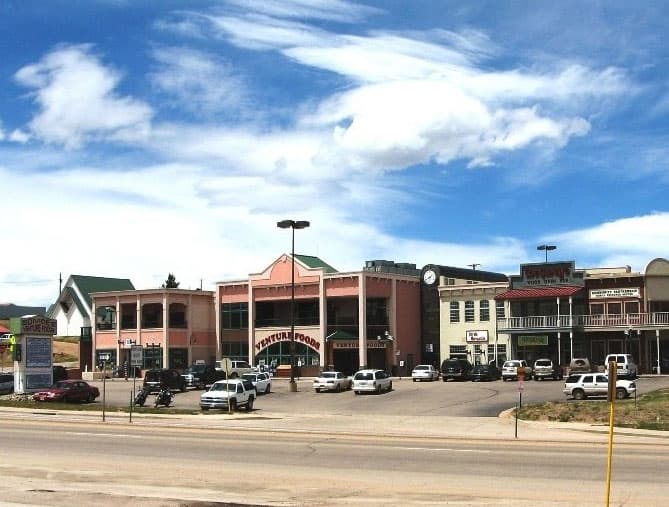 A strip center with multiple architectural styles sits under a blue sky with white, whispy clouds. The parking lot is mainly full and the main building, Ventura Foods is two stories tall and painted a pink tone.