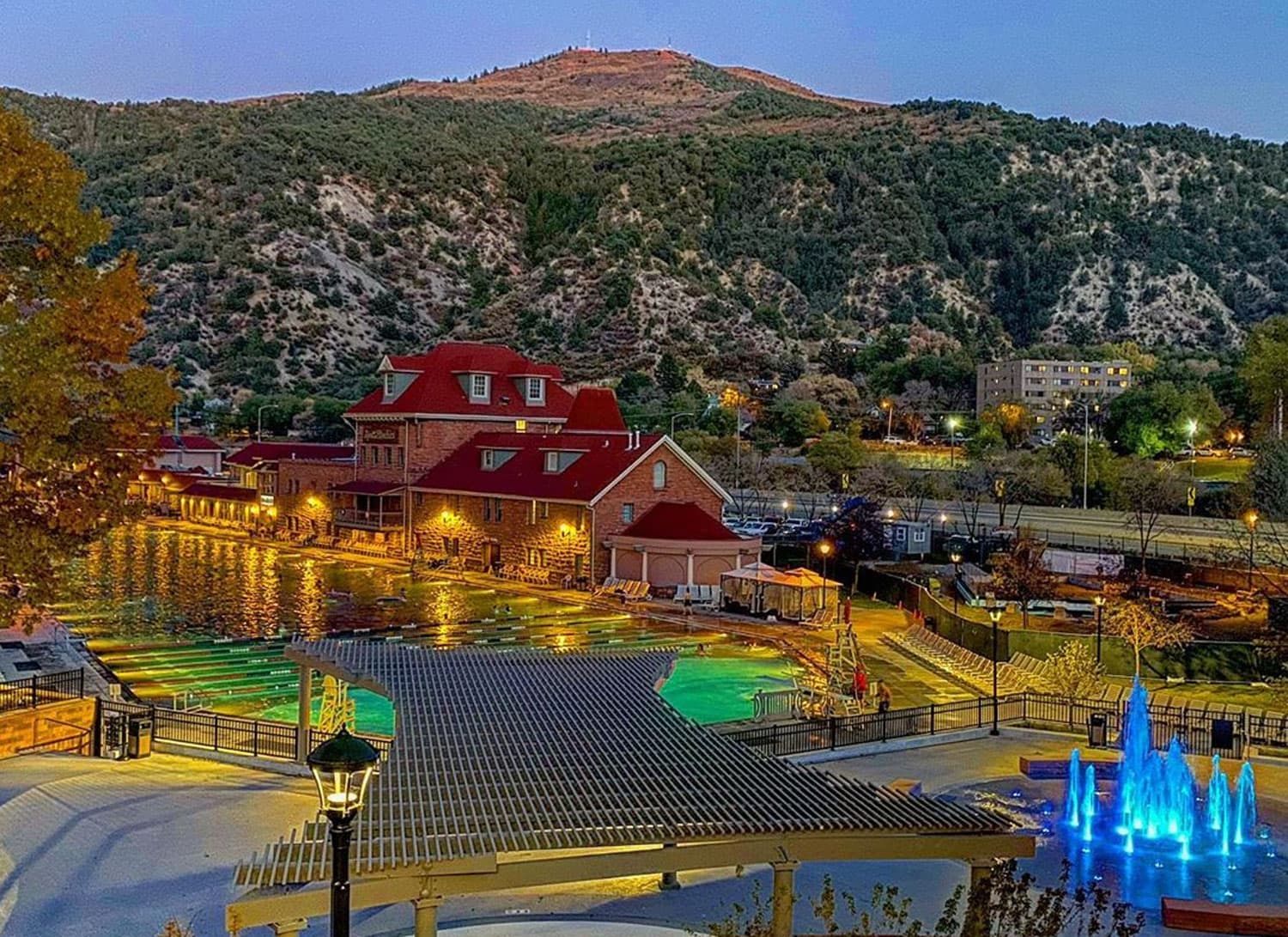 A red-brick building with a red roof sits at the base of a mountain in Glenwood Springs, Colorado. A large hot spring pool is lit with lights and glows blue-green.