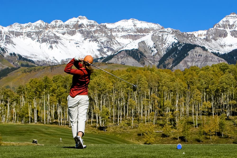 A woman in a bright red top swings for the mountain tops — literally! We can see the jaggest snowcapped peaks of Telluride in the distance. Beneath those, a stand of aspen trees is starting to changes to fall's yellow leaves.