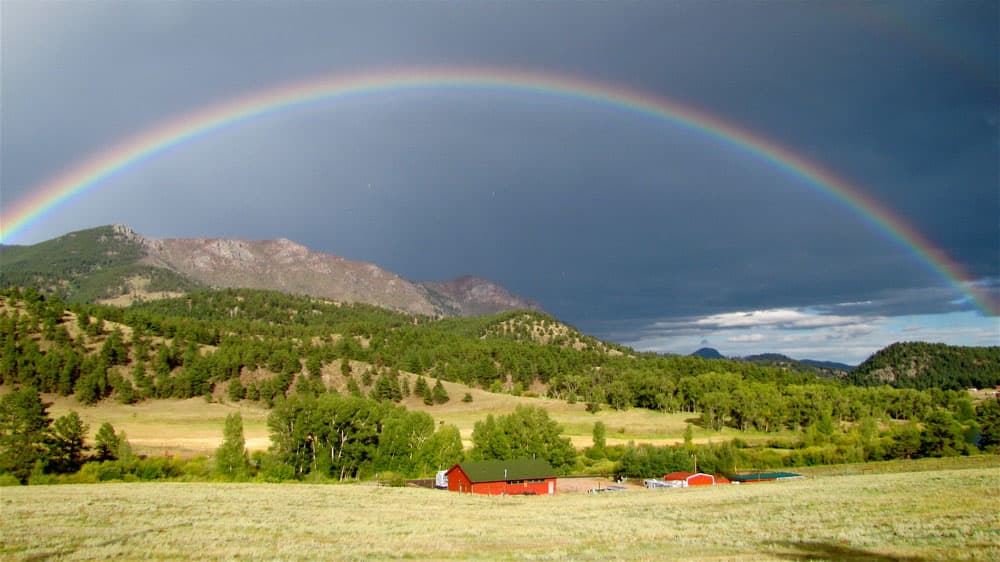A dark stormy sky with a giant rainbow sits above a Rocky Mountain range with green trees and below that a valley of grasses and red barns.