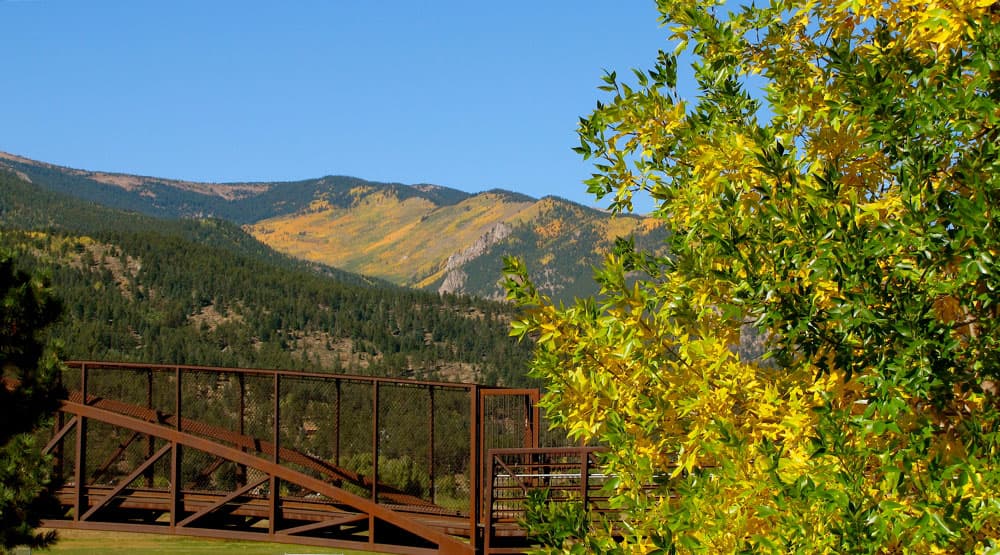A pedestrian metal bridge sits on the left of the image, to the right yellow and green leaves are on a tree. In the distance blue sky meets mountains that are slowly becoming covered in golden hues.