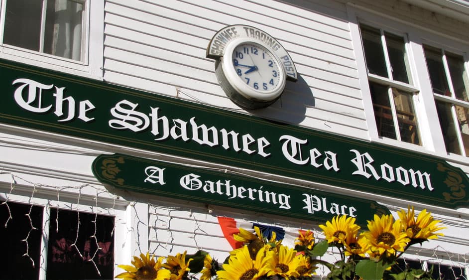A white-wood building with a clock has a sign in old English font that reads "The Shawnee Tea Room A Gathering Place."