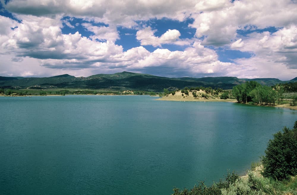 The blue-green hues of the lake sit under the blue sky with white clouds. In the distance green rolling hills lead toward the water.