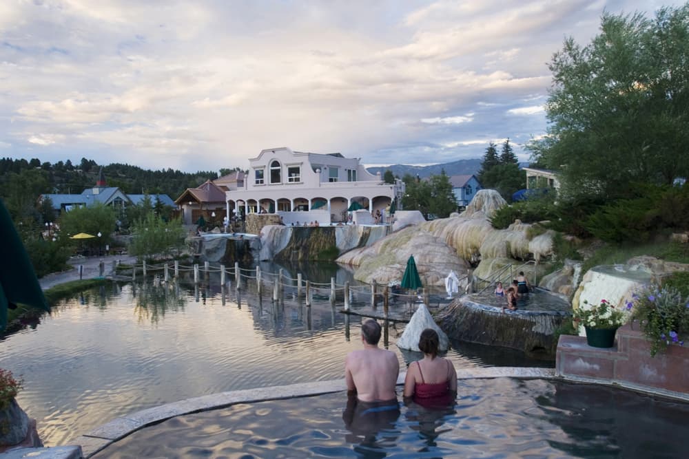 A couple soaks in a hot spring at The Springs Resort & Spa. They are overlooking a bigger pool of water with a white-cloud sky.