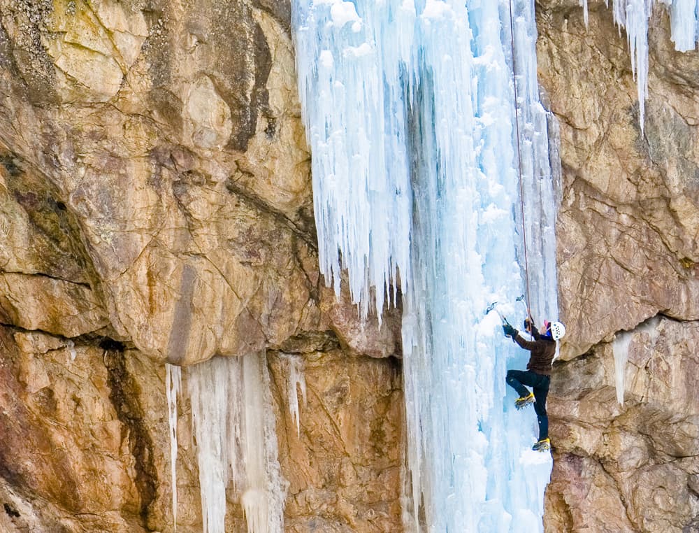 A climber in black scales a section of ice wall using ice picks and spiked boots near Ouray, Colorado.