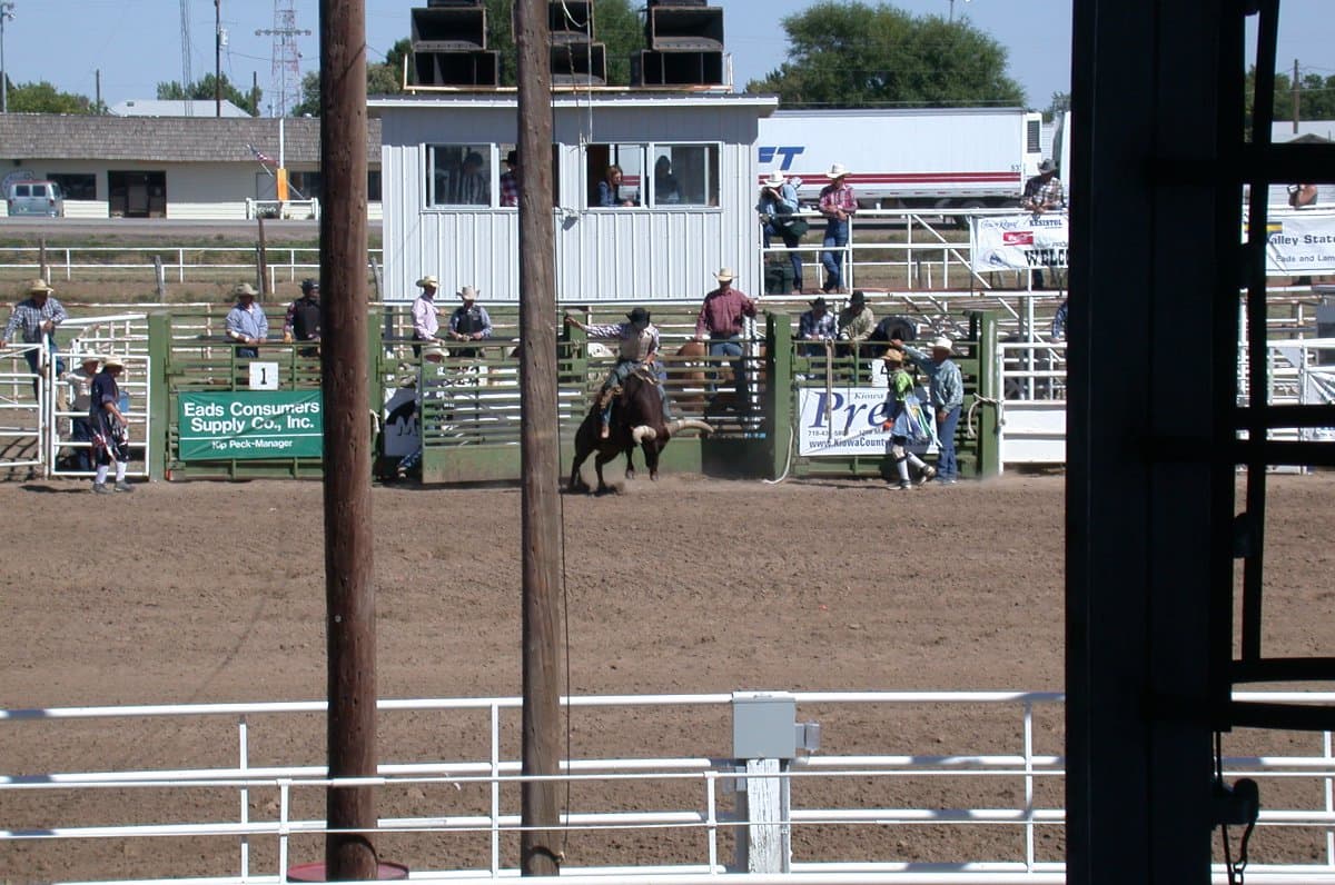 Through the the stands, a bull rider leaves the paddock. There are other people behind in the paddocks and to the right and left of the fences at a rodeo in Kiowa.