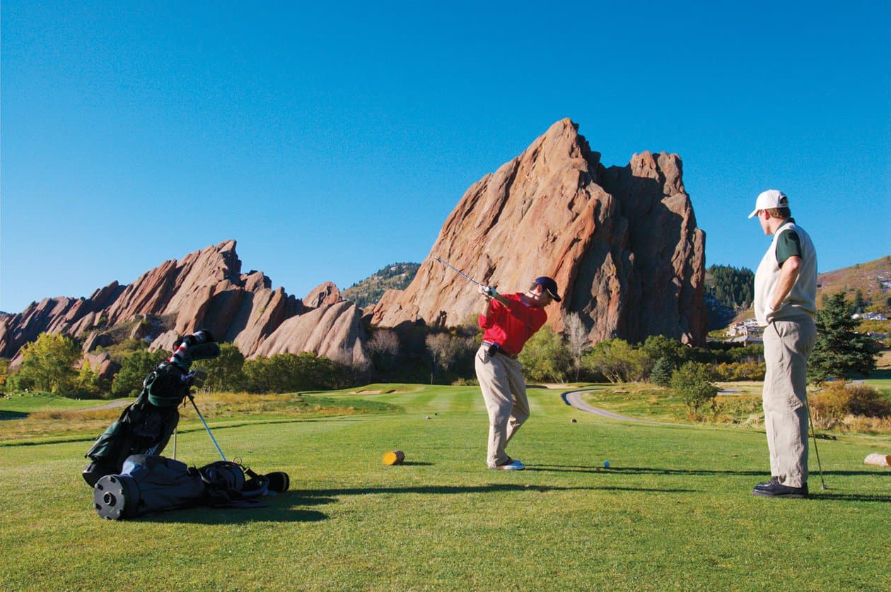 A giant slab of red rock shoots out of the ground surrounded by the golf course's greens. One man watches as another takes a swing. Their golf clubs sit in the foreground. The sky is impossibly blue against the red rocks.