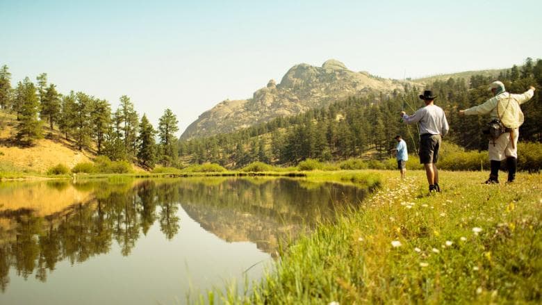 Three people fish on the green-yellow, grass-covered riverbank. The river is glassy, reflecting the evergreen trees and mountain in the background. The light-blue sky is hazy and the image has a yellow hue.