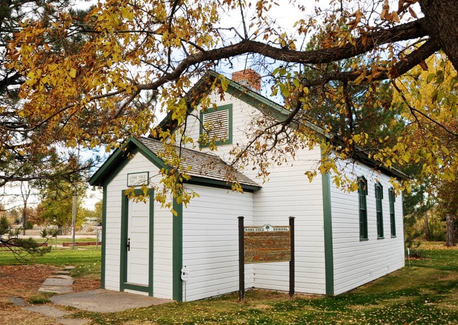 A white-wood sided building with green trim sits on green grass with golden-leafed trees surrounding it.