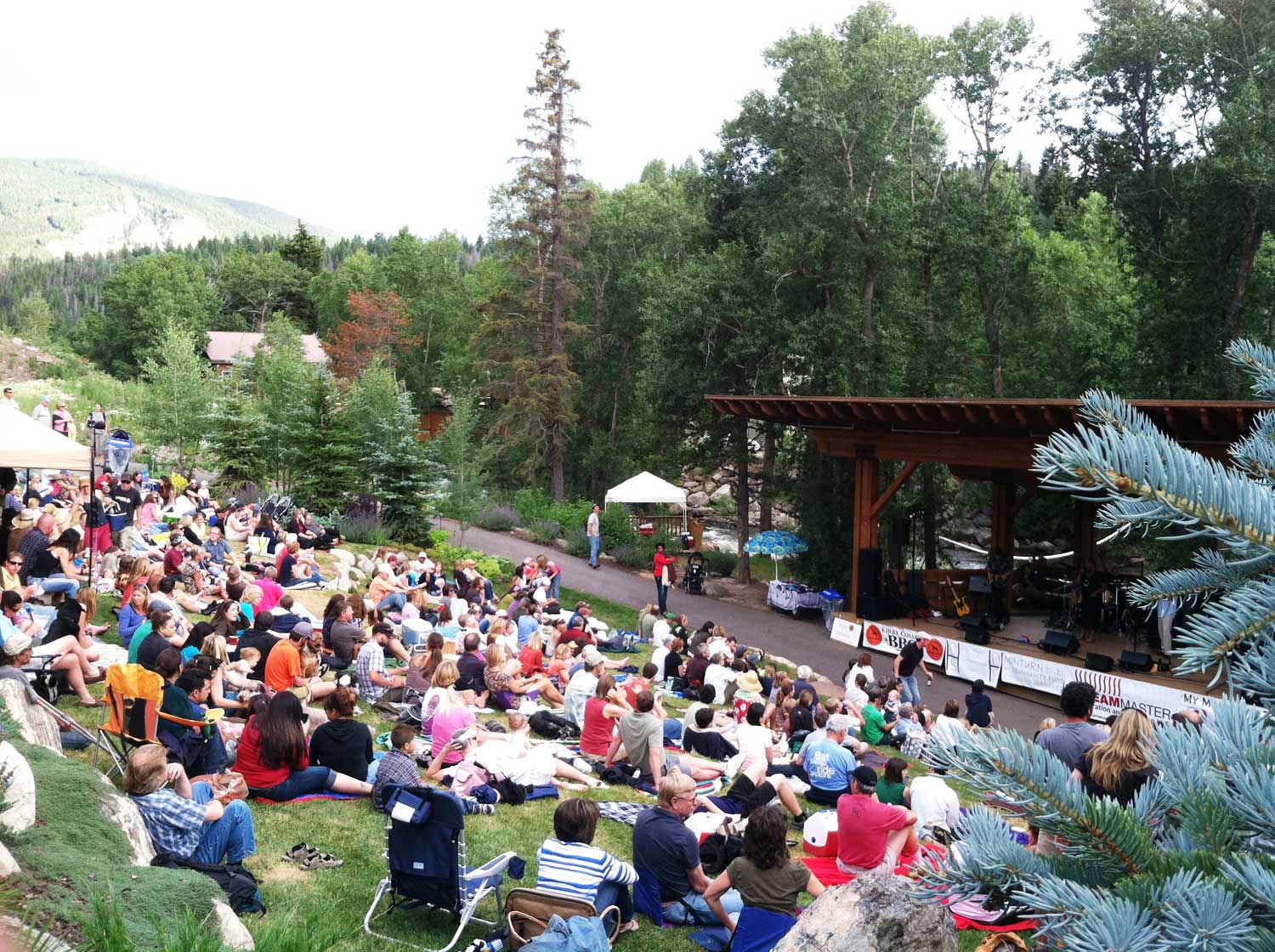 A performance takes place along the river in Minturn. People sit on the grassy slope across the sidewalk from the wooden stage. There's evergreen trees in the background.