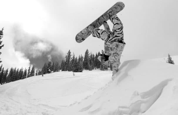 A black-and-white image of a snowboarder doing a handstand in a snow dune with evergreen trees in the background.