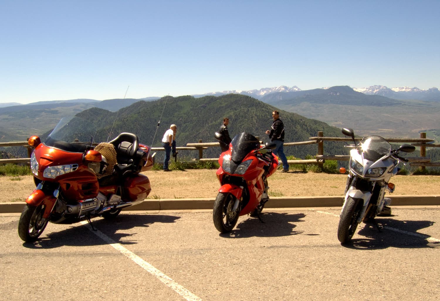 Three motorcycles stand empty in a parking lot overlooking Colorado's San Juan Mountains. Three riders send by the wood-pole fence and admire the scenic view.