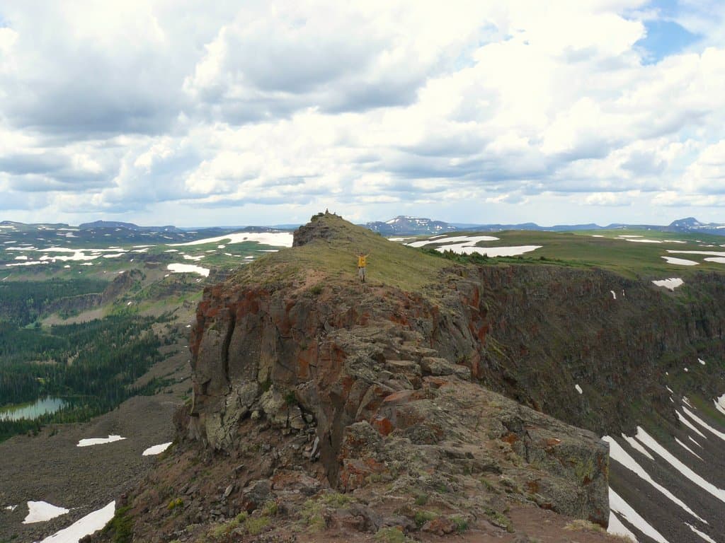 A person hikes Devil's Causeway's narrow trail, rocky gray trail on a cloud-filled summer's day. In the background white snow can be seen in spots.