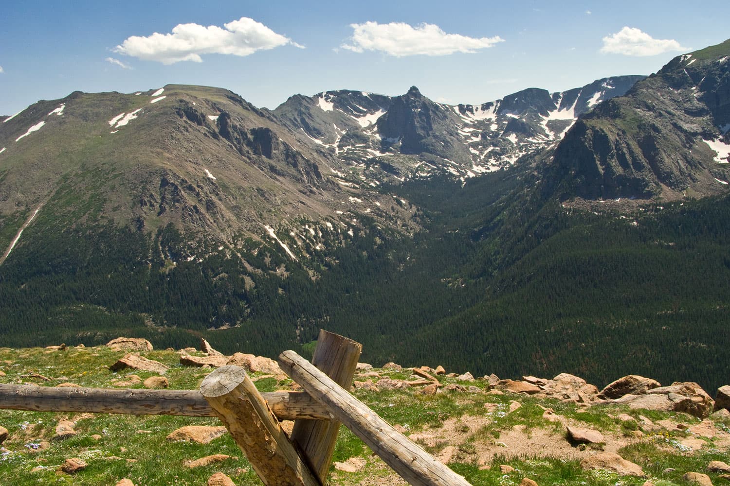 A view taken from an observation point of the Rocky Mountains with some of the stone peaks covered in snow. The sky is a hazy blue with three white clouds. There is green grass with rocks in front of the wooden fence at the observation point.