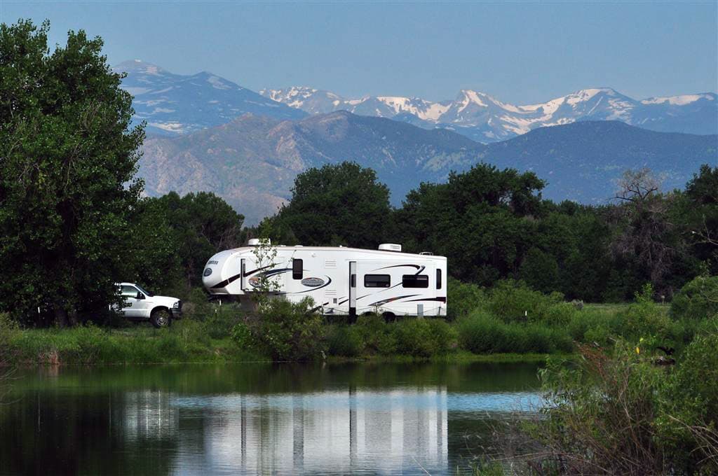 A white RV sits in the middle of a green grass and trees. It's sitting in front of a lake that's reflecting its image. In the background there is a snow-capped mountain range.
