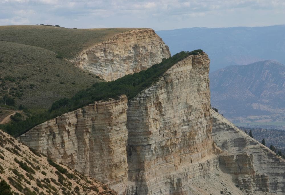 A peak of tan sandstone with a sheer cliff face with the top covered in green grass sits on the left side of the image with hazy mountains in the background.