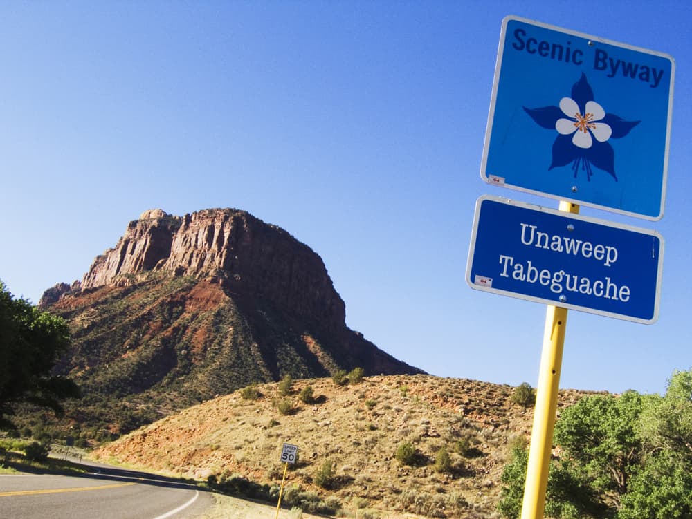 A butte sits along the road with a yellow-grass hill. On the right a square. metal sign with a flower on it says Scenic Byway. On the same sign pole, beneath that it is another metal sign that says "Unaweep Tabeguache."
