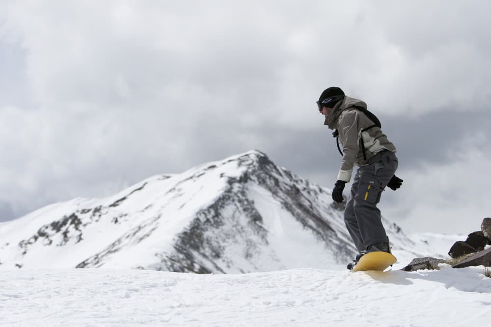 A grey-clothes-wearing snowboarder boards on a yellow snowboard with a snow-covered mountain peak in the background.