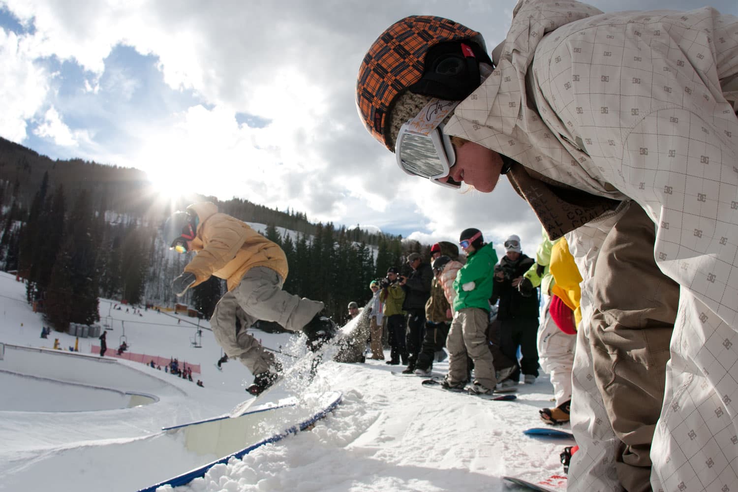 A gathering of snowboarders in helmets and googles waits their turn to hit the halfpipe as the sun sets behind the mountain