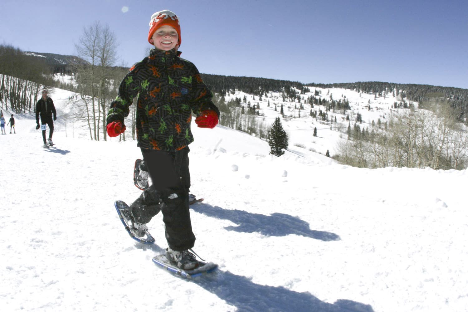 Child snowshoeing on a blue sky winter day. Small trees with no leaves dot the background and a four other people are snowshoeing in the background.