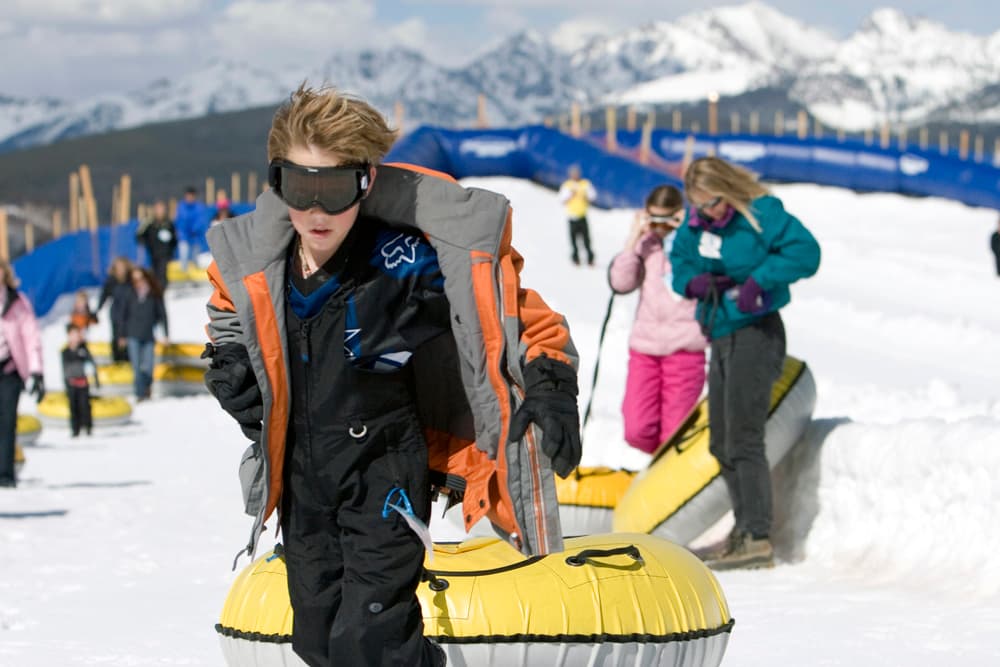 A boy tows his yellow tube on a rope up a hill to get ready to head down it again; several other riders are lined up behind him