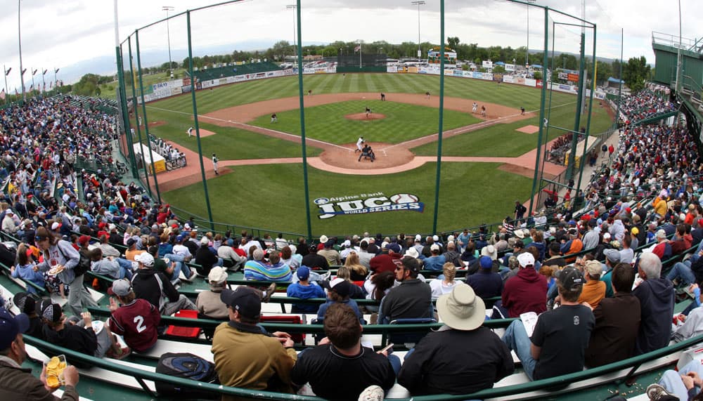 A view from a high seat behind the catcher, shows a modern stadium and crowd at Junior College World Series in Grand Junction, Colorado.