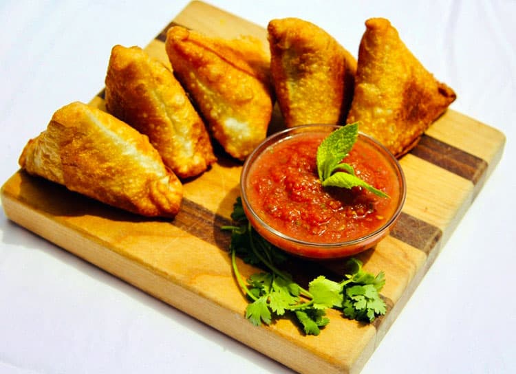 Five crispy samosas rest on a wooden serving board at Susan's Samosas in Colorado. They are served with a small glass dish of a tomato-based sauce.