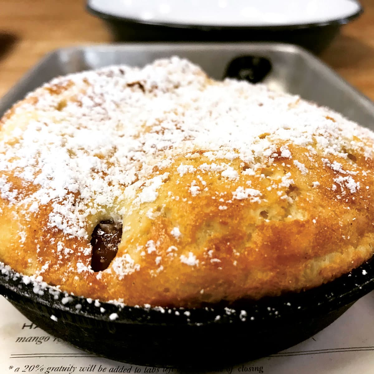 A closeup view shows a fresh pastry cooked to a golden-brown perfection and topped with sweet powdered sugar from Acreage in Colorado. A strategic hole reveals a cherry jam filling.