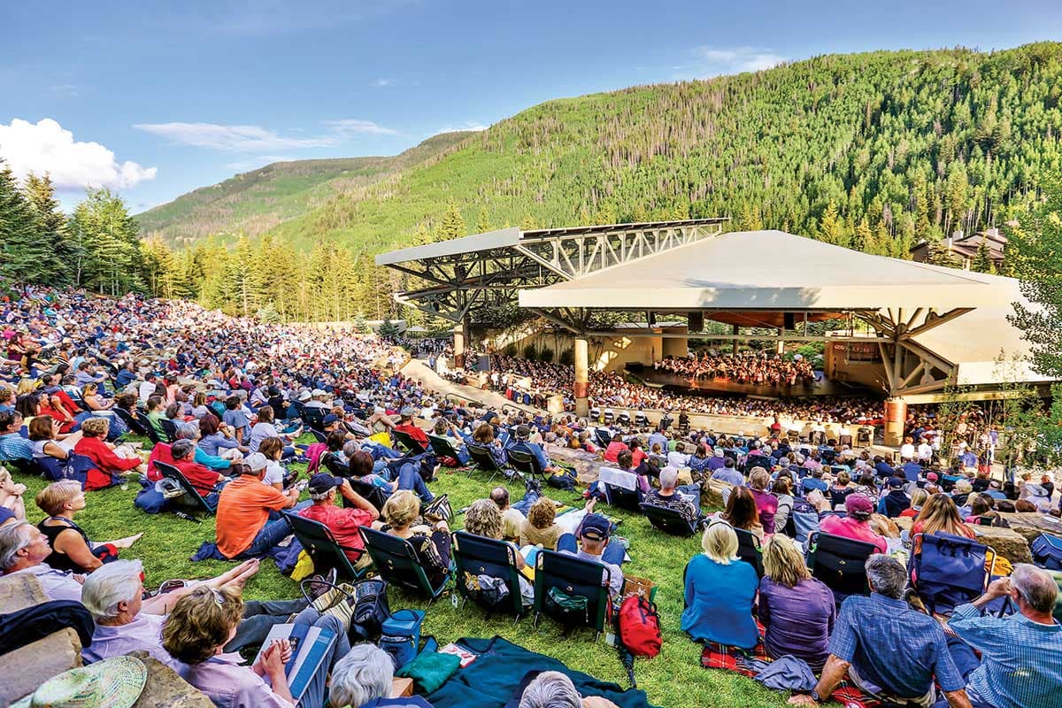 Groups of music fans sit on lawn chairs and blankets facing an amphitheater in Vail, Colorado, and listening to a performance as part of the Bravo! Vail Valley Music Festival.