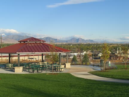 A red-roofed picnic shelter sits near a sidewalk and on top of bright-green grass with the Front Range and trees in the background. The sky is blue with barely any clouds.