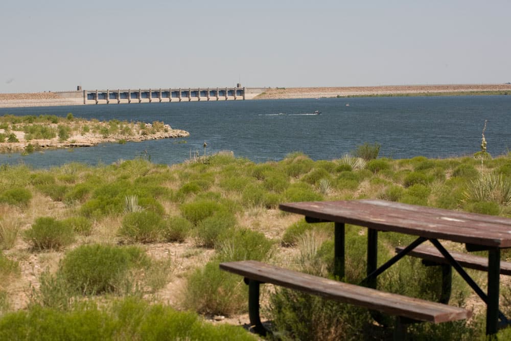 On a summer's day the damn at John Martin Reservoir State Park is in the background with blue water sitting in front of it. In the foreground, a picnic table sits on top of grey and yellow grass shrubs.