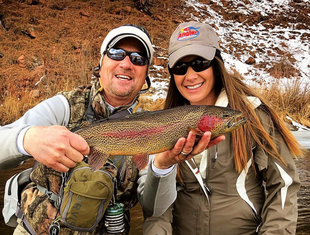 Two wintertime anglers smile and pose with a recently caught freshwater on a river in Colorado. The fish is yellow with brown speckles a red stripe down its side and a blush spot around its gills.