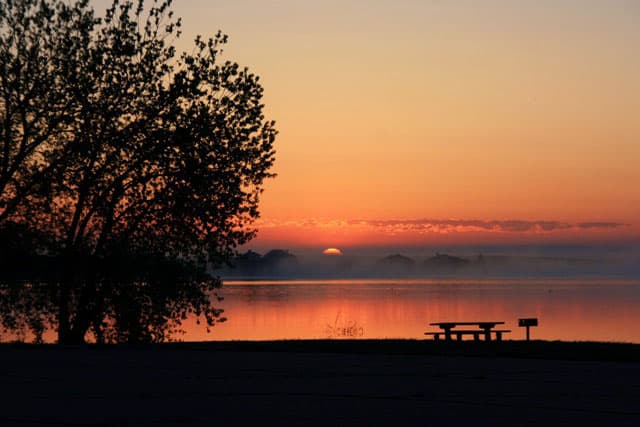 The sun is setting over a the lake at Boyd Lake State Park. A fog is settling in over the lake and a massive tree is to the left of a picnic table and grill. The sky is orange, yellow, pink and a bit blue.