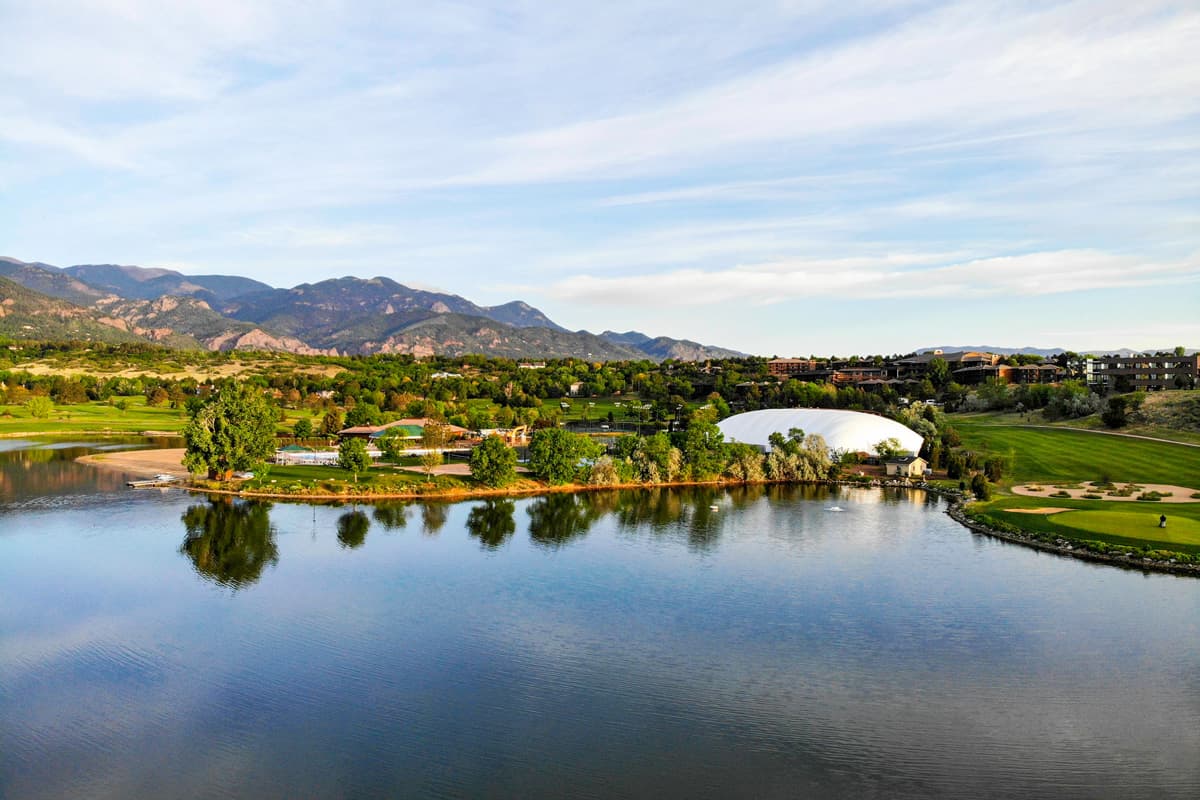 Bluebird skies with wisps of clouds are reflected in the calm waters near Cheyenne Mountain Resort. There is a golf course by the edge of the water and the resort area looks lush with many mature trees.