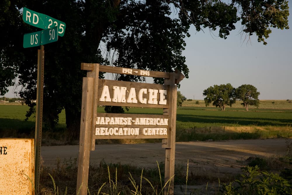 On a summer's day, there is a wooden sign in the middle that reads "1942–1945 Amache Japanese-American Relocation Center." To the left of that is a green metal street sign, with the top rectangle saying "RD 235" and the bottom rectangle saying "US 50." In the background there are green trees, green fields and a brown dirt road.