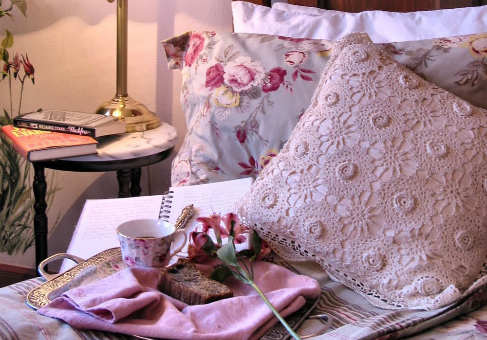 A breakfast tray with a slice of fruit cake, a flowered tea cup and pink flower sits by an open journal with elegant cursive writing on a pillow-ladened bed in Denver, Colorado.