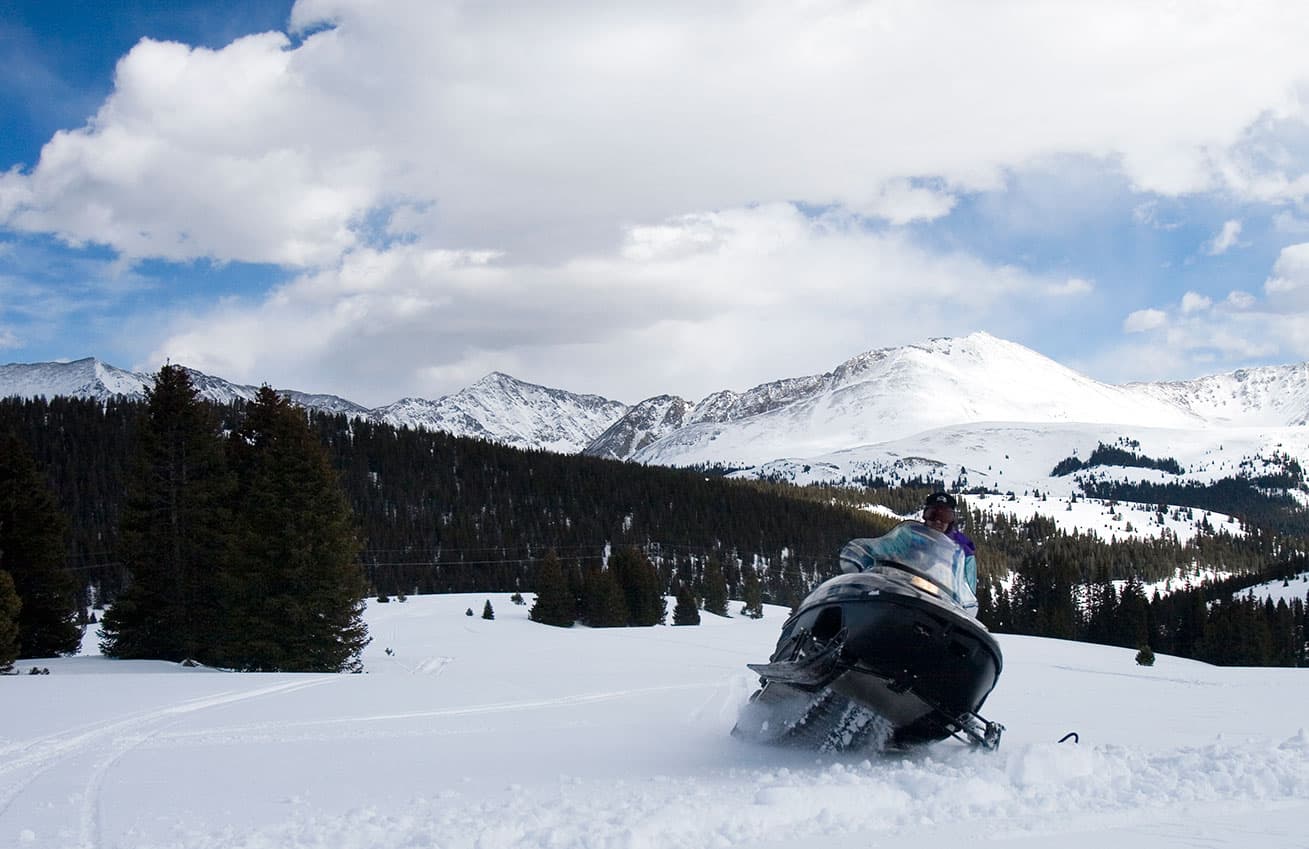 Snowmobilers ride through fields of fresh powder near Vail Colorado. They are surrounded by pine trees and in the distance are towering snow-covered peaks.