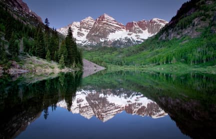 The two peaks of the Maroon Bells are reflected in the lake below. The sky is blue and green forest surrounds the lake. The peaks are dusted in snow.
