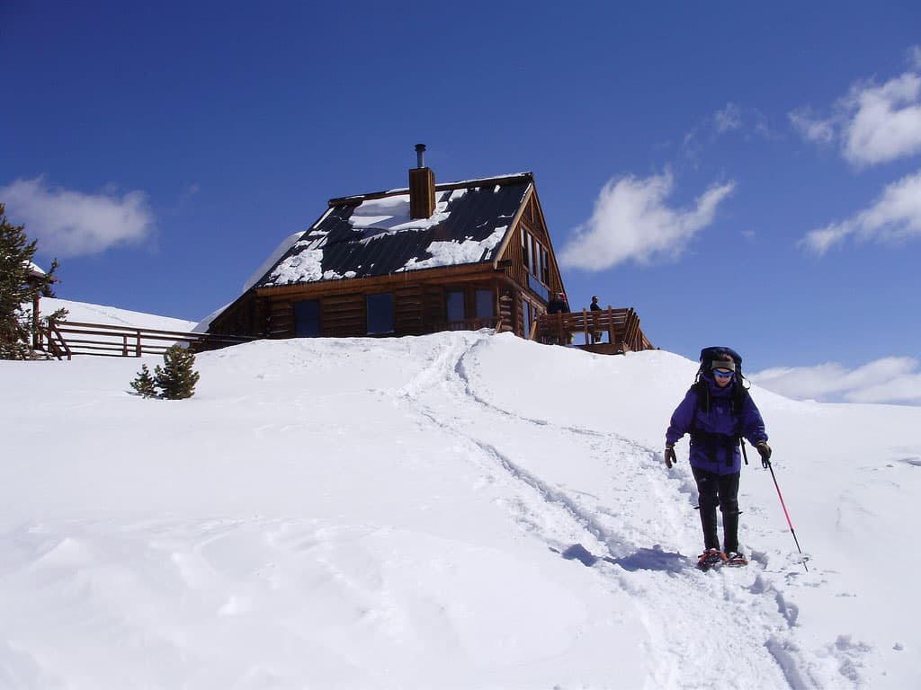 A snowshoer makes tracks past a snowy cabin in the background