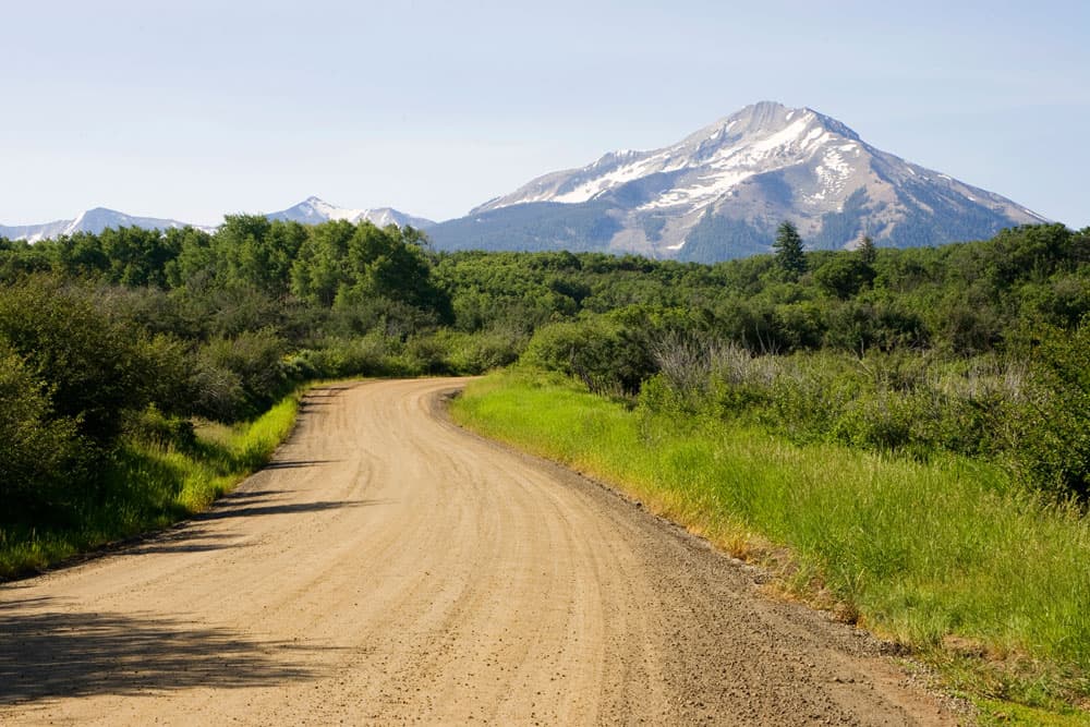 A dirt road is surrounded by green grass and green bushes on a hazy day with snow-covered stone mountains on a route between Somerset and Crested Butte.