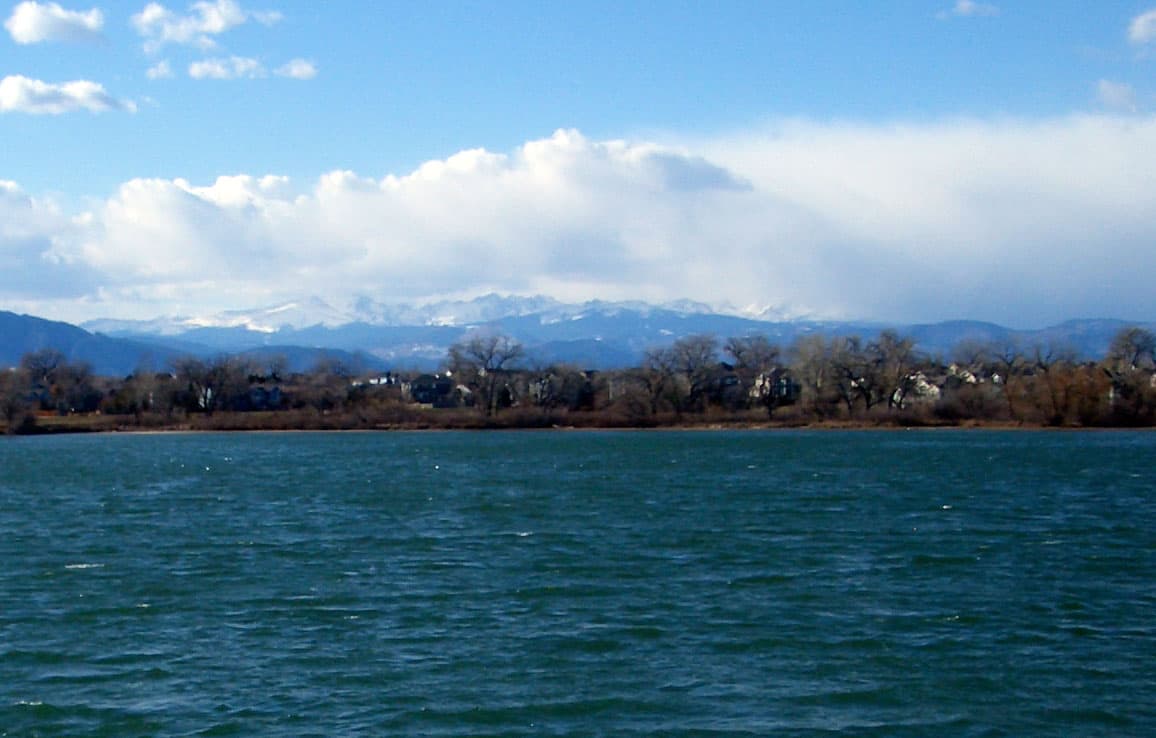 The blue-green water is rippling on Lafayette's Waneka Lake. In the distance, the snow-capped Front Range sits with blue skies and puffy white clouds.
