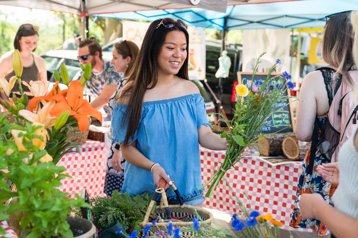 At a market in Boulder, Colorado, a person holds a woven basket in one hand and in the other a bright bouquet with yellow, lavender-purple and indigo-blue flowers.