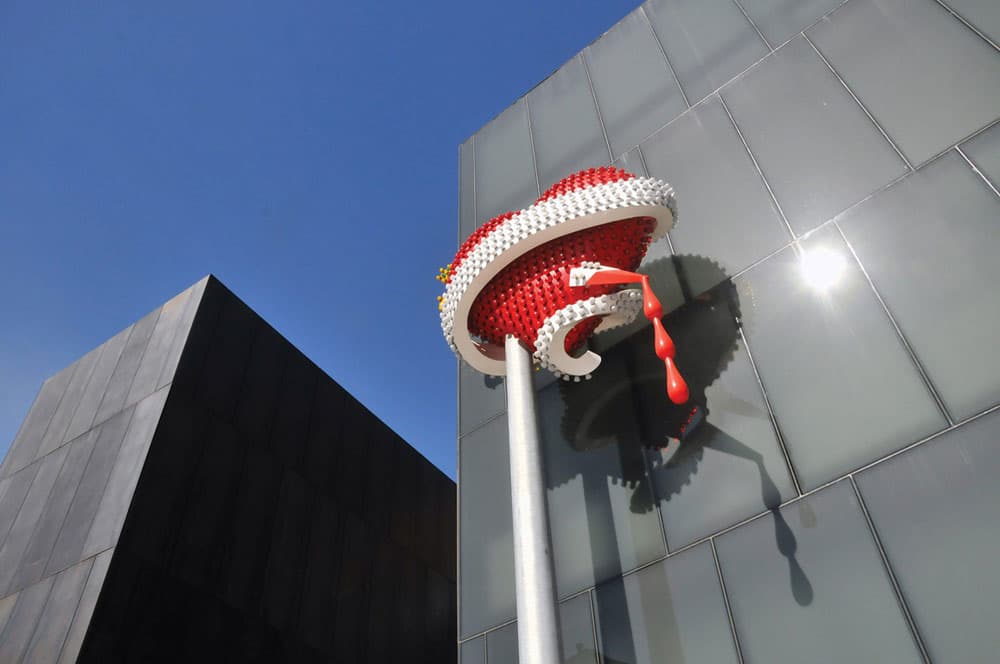 A statue/art piece on the outside of the Museum of Contemporary Art in Denver, Colorado. The art piece, named oxic Schizophrenia / Hyper Version by Tim Novle and Sue Webster, is a large, bedazzled, red heart with blood dripping out of it because of a large knife stabbiing through the other side. The sky above the art is deep blue.