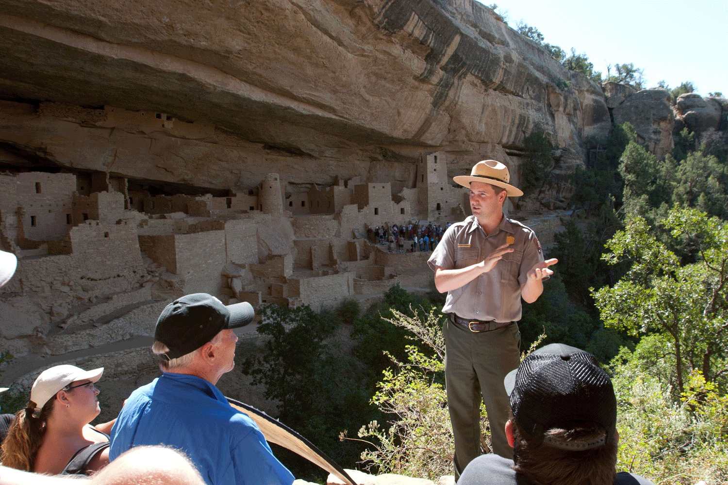 A park ranger speaks to a tour group at Mesa Verde National Park in Colorado. Behind the ranger, the cliff dwellings are shaded from the warm summer sun.