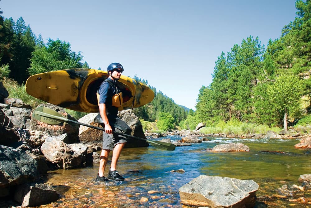 On a blue-sky summer day, surrounded by green pine trees, a kayaker stands with their feet in the water, wearing a helmet, scouts the Cache la Poudre River while holding their kayak over their shoulder.